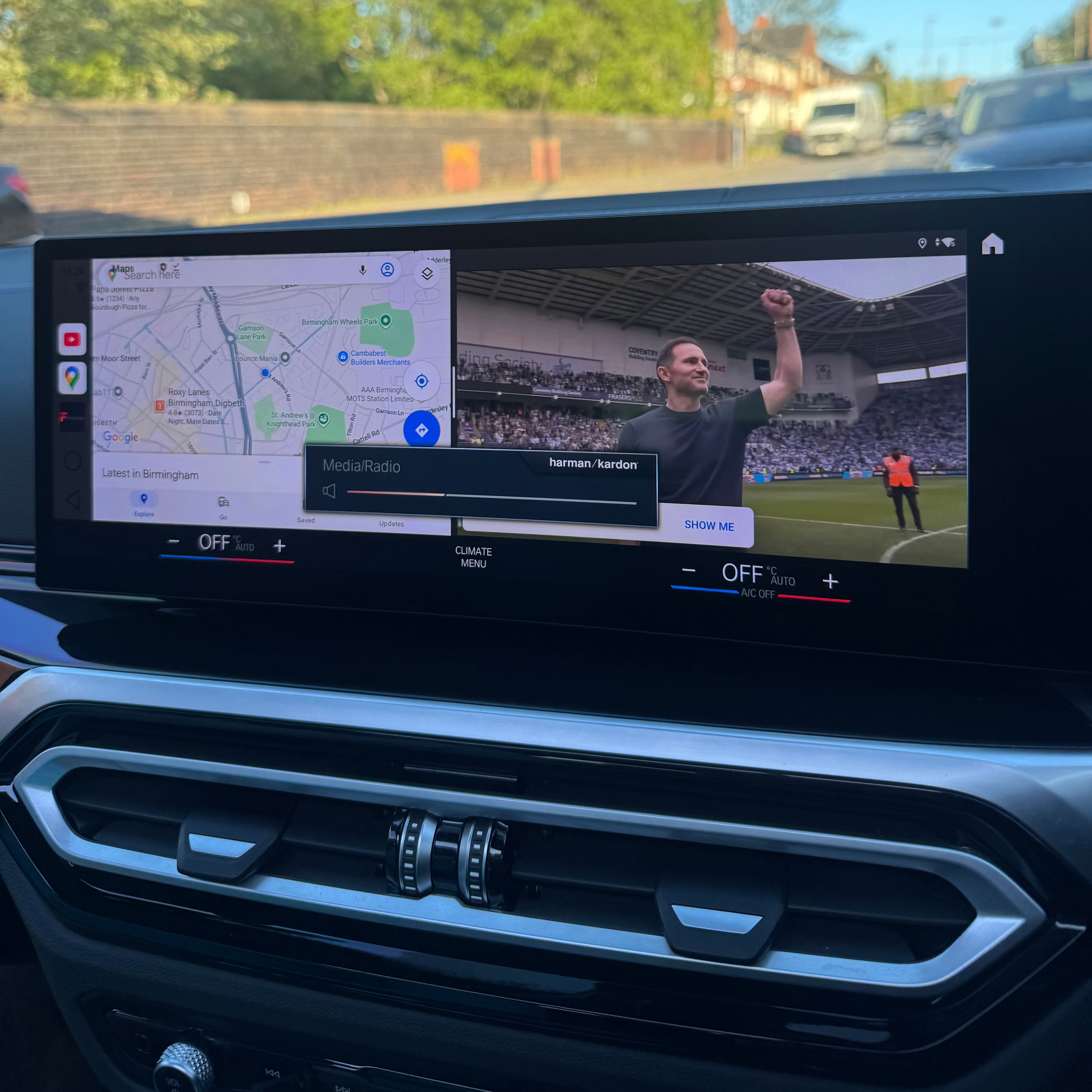 Car dashboard with a navigation screen displaying a map and a video of a man in a stadium.
